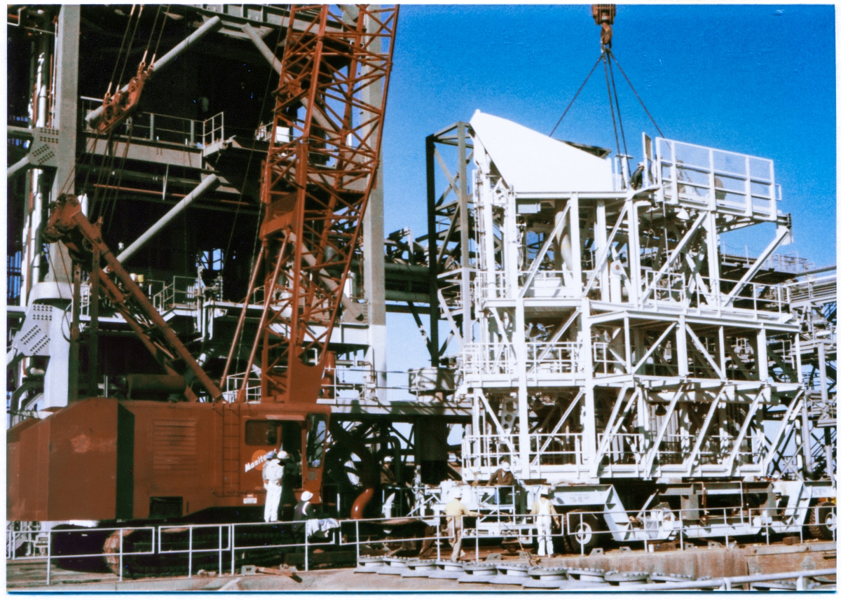 Image 121. Viewed from across the Flame Trench, beyond the row of Spray Headers (bottom right margin of this image) which run along the crest of the Flame Deflector at Space Shuttle Launch Complex 39-B, Kennedy Space Center, Florida, the Intertank Access Arm structure, which you see in white paint, continues to rest on its special transporter which has been rolled up to the Pad Deck, just in front of the MLP Utilities Interface Platform and the 9099 Building, adjacent to the base of the Fixed Service Structure. Union Ironworkers from Local 808, working for Ivey Steel, can be seen up on top of it, making final leveling adjustments to the four-legged lifting sling which will be used to raise it up to elevation 220'-0”, where it will be bolted on to the massive support framework on the north side of the tower, which had previously been added to the LUT which became the FSS by Wilhoit Steel Erectors, several years previously. Photo by James MacLaren.